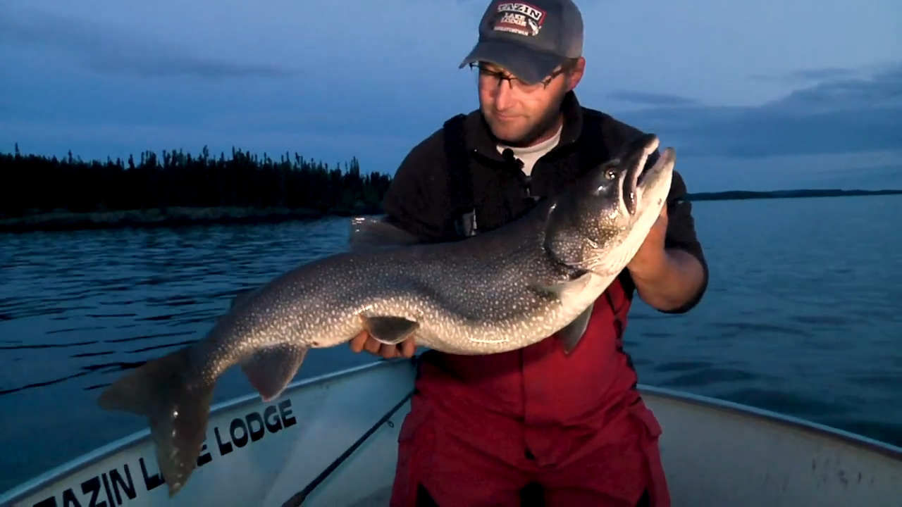 Man holding a large lake trout on a boat at dusk, showcasing an extraordinary fishing achievement