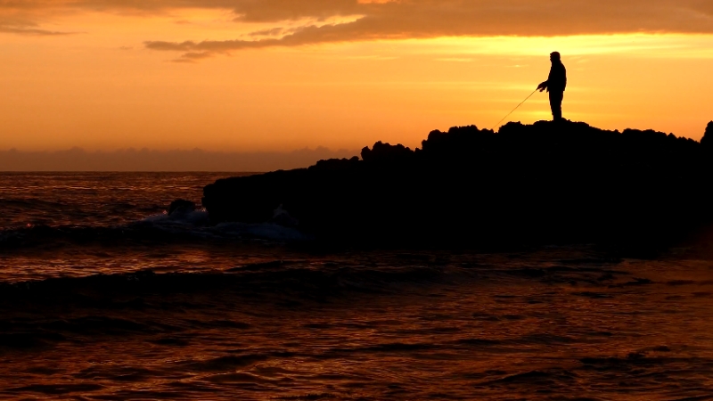 A lone fisherman casting a line into the ocean during sunset