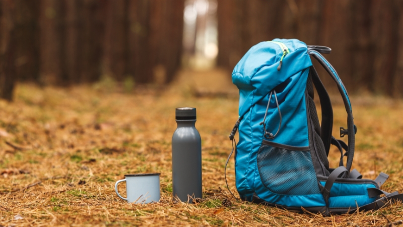 Backpack, bottle, and mug set on a forest path, representing gear essentials for building a strong outdoor brand presence