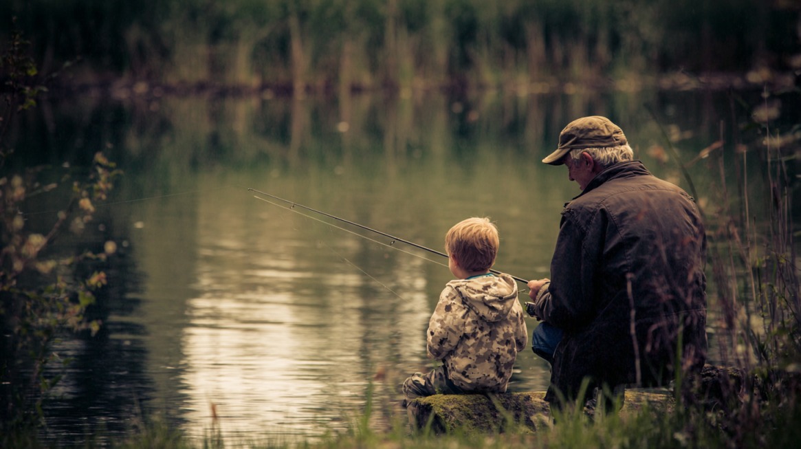 An older man and a young boy sit side by side on the bank of a calm lake, both holding fishing rods and casting lines into the water