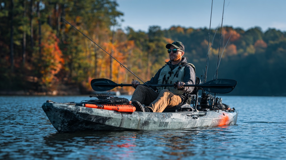 Man fishing from a camo kayak on a calm lake with autumn trees in the background