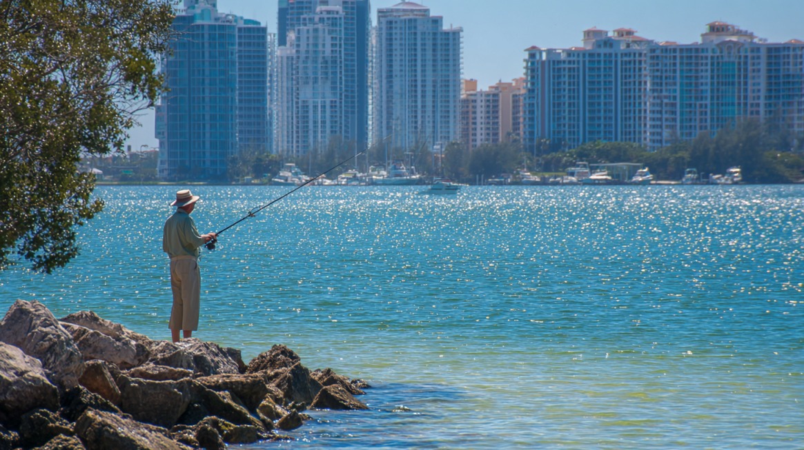 Man fishing from rocks along the shore with high-rise buildings across the water in Sarasota, Florida