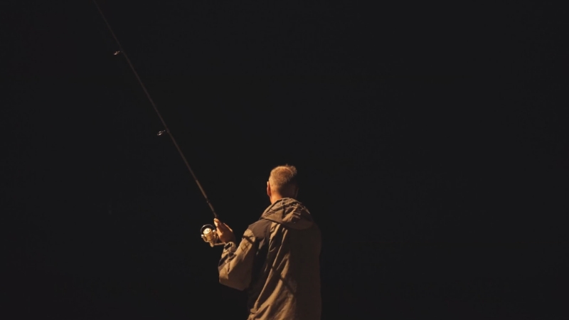 A man stands near dark water holding a fishing rod during night fishing