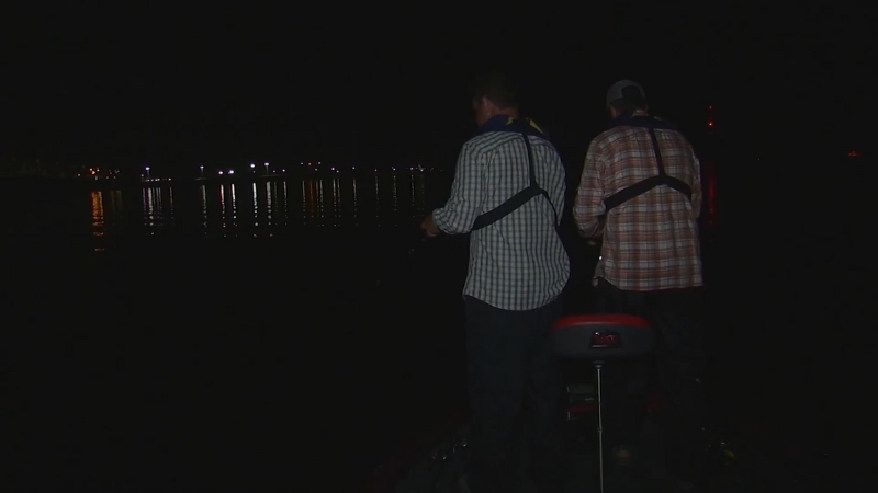 Two anglers stand on a boat fishing in the dark on Kentucky Lake with distant lights reflected on the water