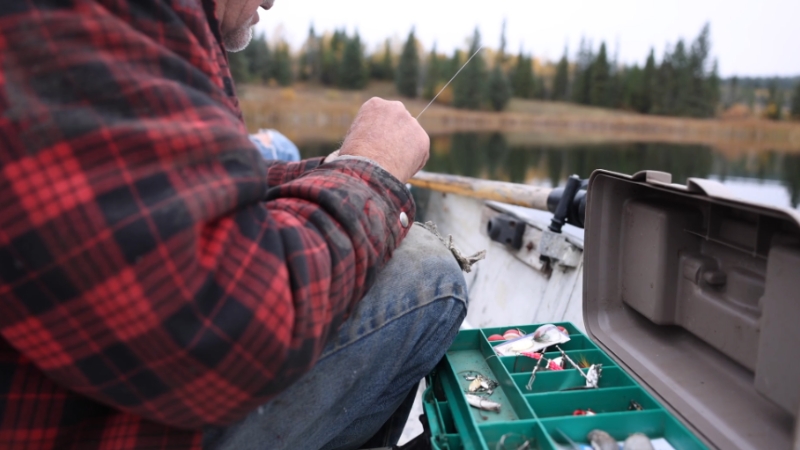An angler sits in a boat choosing lures from a tackle box near a calm lake