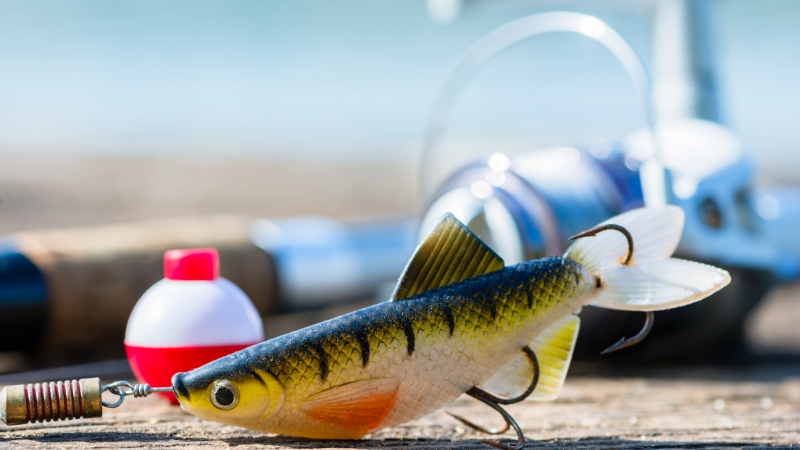A soft plastic fish lure sits on a dock beside a bobber and a spinning reel