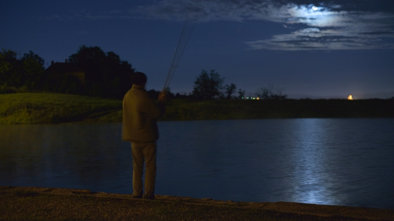 A person stands beside a calm lake at night holding a fishing rod under moonlight during night fishing