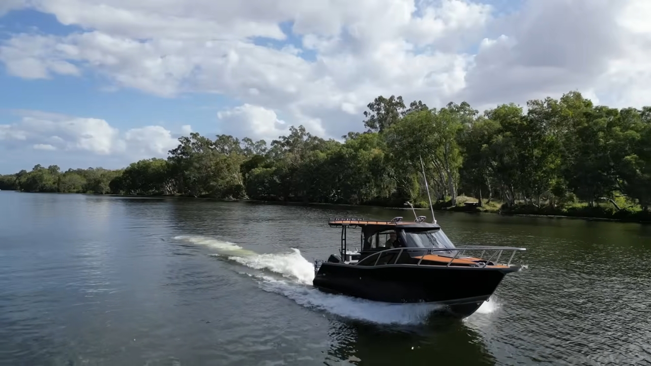 A black aluminum boat rides across a calm river near a tree-lined shore
