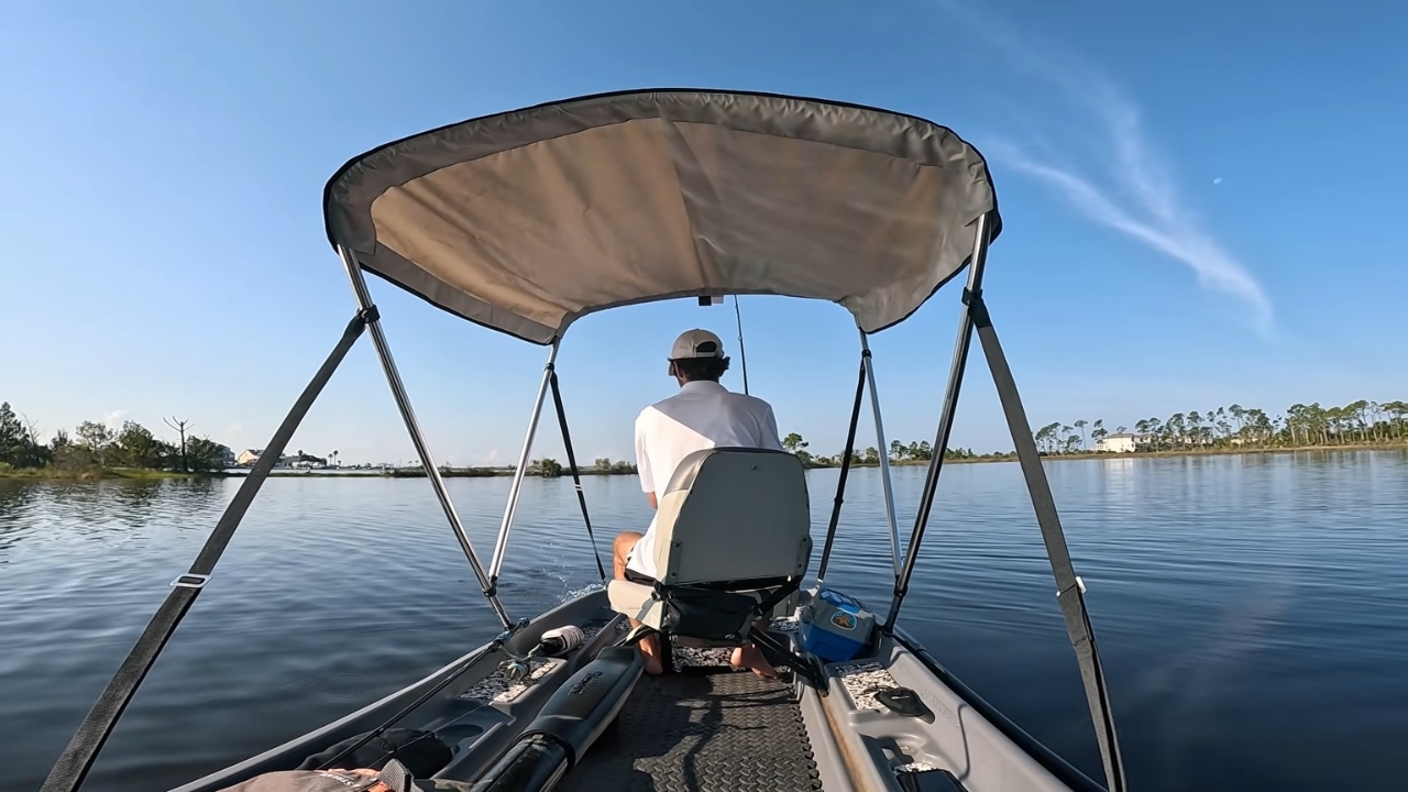 A man sits under a canopy on a small pontoon boat on a calm lake